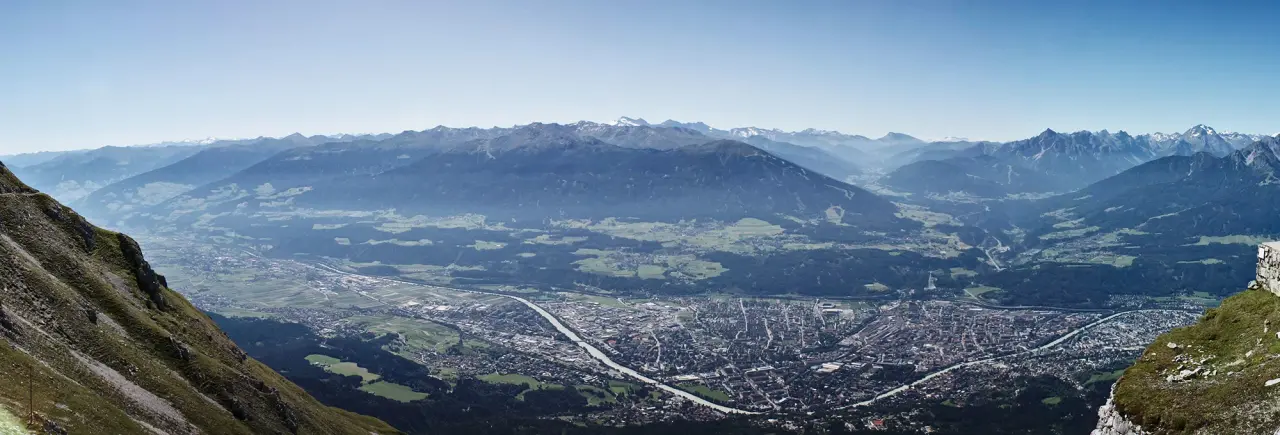 Panoramablick auf das Schweizer Alpen-Tal, der eine weitläufige Stadt inmitten von sattgrünen Feldern und fernen schneebedeckten Bergen unter einem klaren blauen Himmel zeigt.